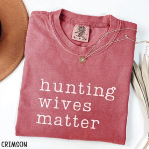 K&ouml;nnte beinhalten: Karmesinrotes T-Shirt mit dem wei&szlig;en Text "hunting wives matter". Das Shirt hat einen Rundhalsausschnitt und ein kleines Etikett in der N&auml;he des Kragens. Eine silberne Halskette mit Anh&auml;nger liegt &uuml;ber dem Shirt. Ein brauner Hut ist oben links zu sehen.