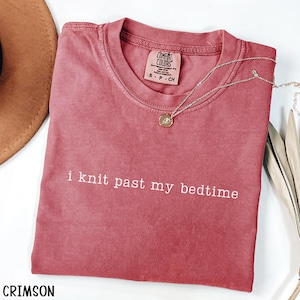 Puede incluir: Camiseta de color carmes&iacute; con el texto blanco "i knit past my bedtime". La camiseta tiene cuello redondo y mangas cortas. Un collar plateado con un colgante redondo est&aacute; drapeado sobre la camiseta. La camiseta est&aacute; etiquetada como "Comfort Colors".