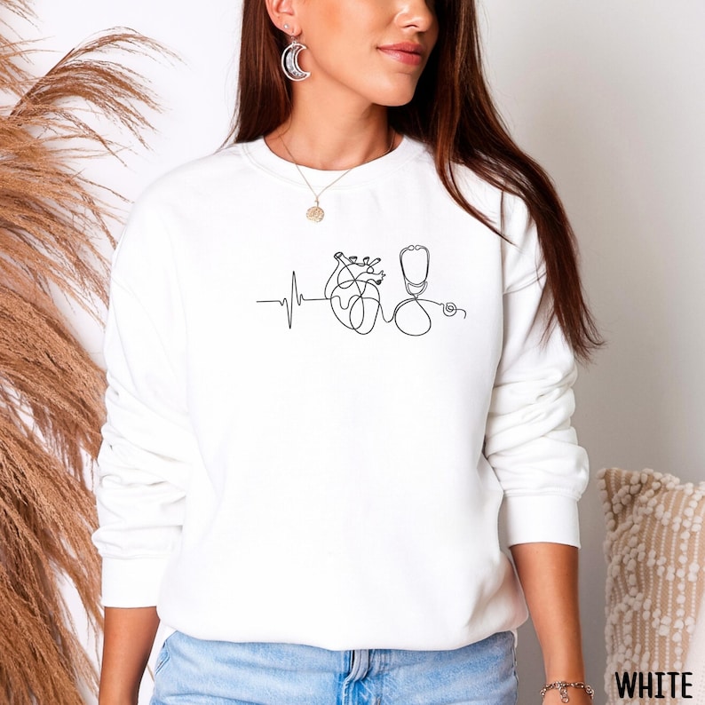 May include: White sweatshirt featuring a minimalist line drawing of a heart, stethoscope, and heartbeat. The design is in black and is centered on the front of the long-sleeved top. The word "WHITE" is printed in white on the bottom right corner.