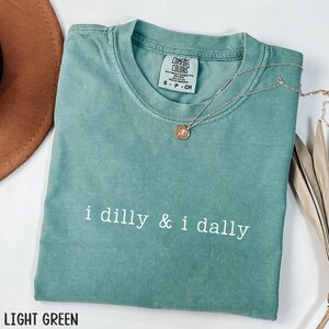 May include: Light green Comfort Colors t-shirt with the phrase "i dilly & i dally" in white. A gold necklace with a pendant rests on the shirt. The shirt is folded, and a brown hat is partially visible.