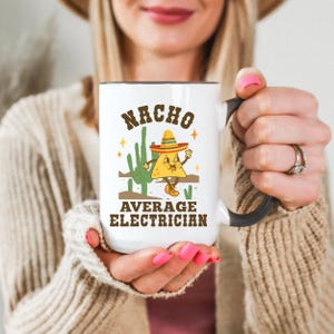 May include: White ceramic mug with a black handle and rim, featuring the text "NACHO AVERAGE ELECTRICIAN" in brown letters. The design includes a cartoon nacho wearing a sombrero and a cactus. The mug is held by a person.