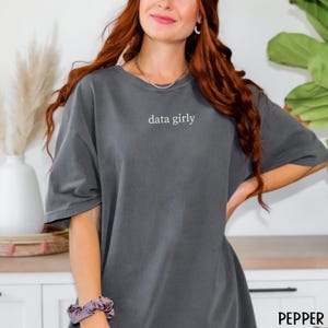 May include: Dark grey t-shirt with the text "data girly" in white. The shirt has short sleeves and a relaxed fit. The word "PEPPER" is printed in the bottom right corner.