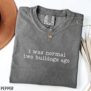 May include: A gray Comfort Colors t-shirt with the text "i was normal two bulldogs ago" in white. A silver necklace with a pendant rests on the shirt. A brown hat is visible in the upper left corner.