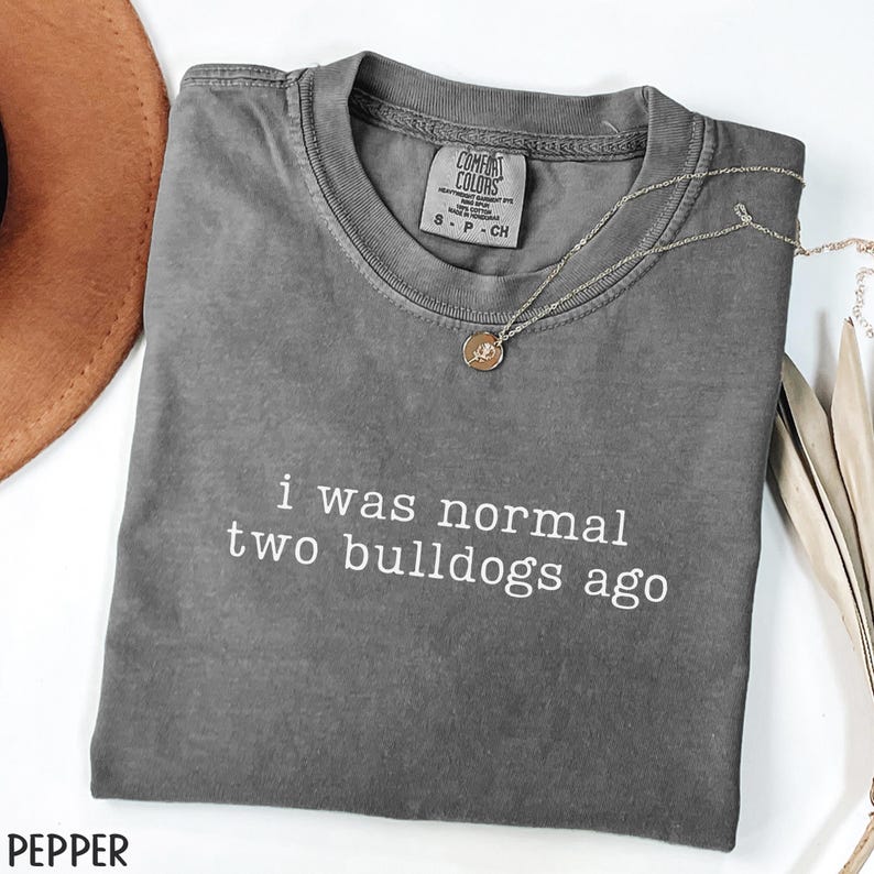 May include: A gray Comfort Colors t-shirt with the text "i was normal two bulldogs ago" in white. A silver necklace with a pendant rests on the shirt. A brown hat is visible in the upper left corner.
