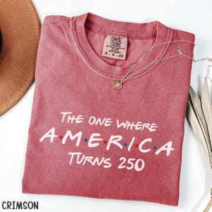 K&ouml;nnte beinhalten: Karmesinrotes T-Shirt mit dem wei&szlig;en Text "The One Where America Turns 250". Das Wort "America" hat rote und blaue Punkte. Eine silberne Halskette mit Anh&auml;nger liegt auf dem Shirt. Ein brauner Hut ist im Hintergrund zu sehen.