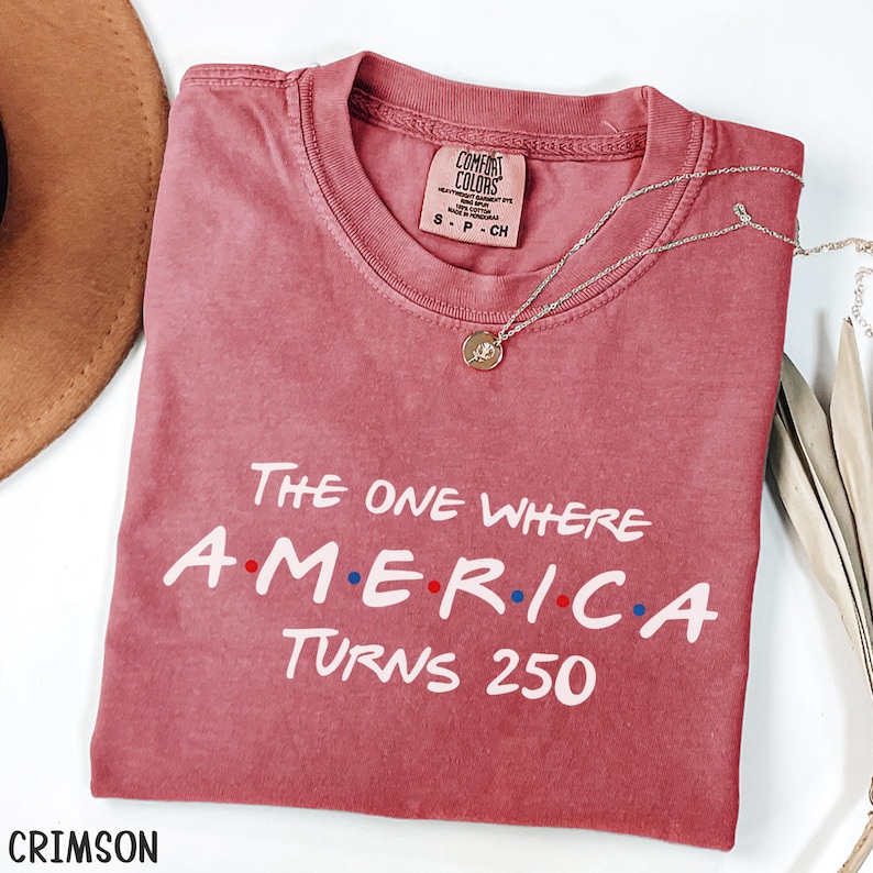 K&ouml;nnte beinhalten: Karmesinrotes T-Shirt mit dem wei&szlig;en Text "The One Where America Turns 250". Das Wort "America" hat rote und blaue Punkte. Eine silberne Halskette mit Anh&auml;nger liegt auf dem Shirt. Ein brauner Hut ist im Hintergrund zu sehen.