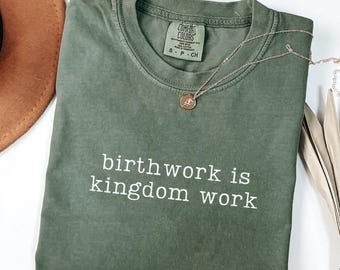 May include: A moss green t-shirt with the text "birthwork is kingdom work" in white. A gold necklace with a pendant rests on the shirt. The shirt is folded and displayed with a brown hat and dried leaves.