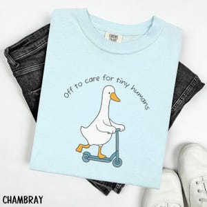 May include: Light blue t-shirt with a graphic of a white goose riding a blue scooter. The text "off to care for tiny humans" is arched above the goose. The shirt is paired with black jeans and white sneakers.