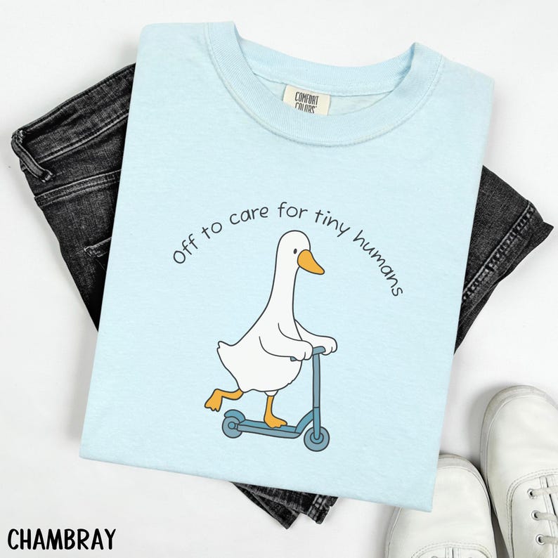 May include: Light blue t-shirt with a graphic of a white goose riding a blue scooter. The text "off to care for tiny humans" is arched above the goose. The shirt is paired with black jeans and white sneakers.