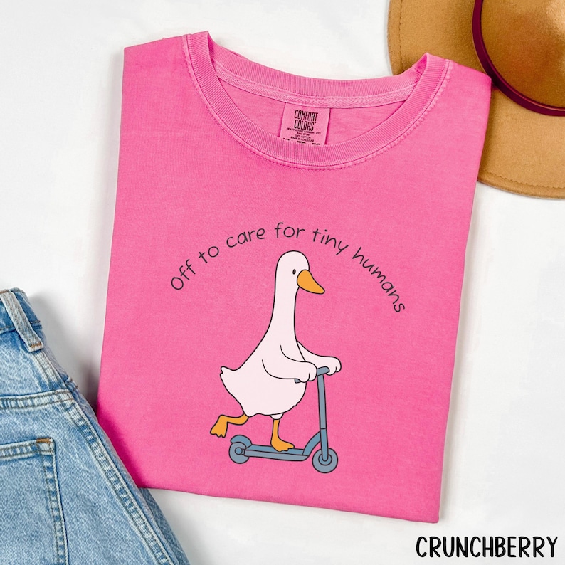 May include: A bright pink t-shirt with the text "Off to care for tiny humans" arched above a cartoon goose riding a blue scooter. The goose is white with a yellow beak and feet. The brand name "CRUNCHBERRY" is at the bottom.