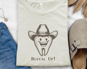 May include: A light beige t-shirt with a cartoon drawing of a tooth wearing a cowboy hat. The tooth is smiling and the text "Buccal Up!" is printed below the tooth.