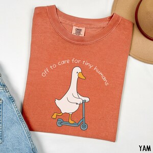 May include: Rust-colored Comfort Colors t-shirt with a white cartoon goose riding a blue scooter. The text "Off to care for tiny humans" is arched above the goose. A tan hat and denim overalls are partially visible.