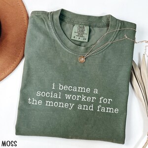 May include: A moss green Comfort Colors t-shirt with the text "i became a social worker for the money and fame" in white. A gold necklace with a pendant is on the shirt. A brown hat is partially visible in the upper left corner.