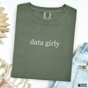 May include: A moss green t-shirt with the words "data girly" in white script. The shirt is folded and has a Comfort Colors tag. A pair of blue jeans is visible in the bottom right corner.