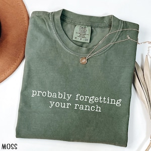 May include: A moss green t-shirt with the phrase "probably forgetting your ranch" printed in white. A gold-colored necklace with a pendant is on the shirt. A brown hat is partially visible in the upper left corner.