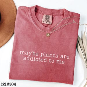 Może przedstawiać: Koszulka w kolorze karmazynowym z białym napisem "maybe plants are addicted to me". Srebrny naszyjnik z małą zawieszką spoczywa na koszulce. Koszulka ma okrągły dekolt i metkę z napisem "Comfort Colors".
