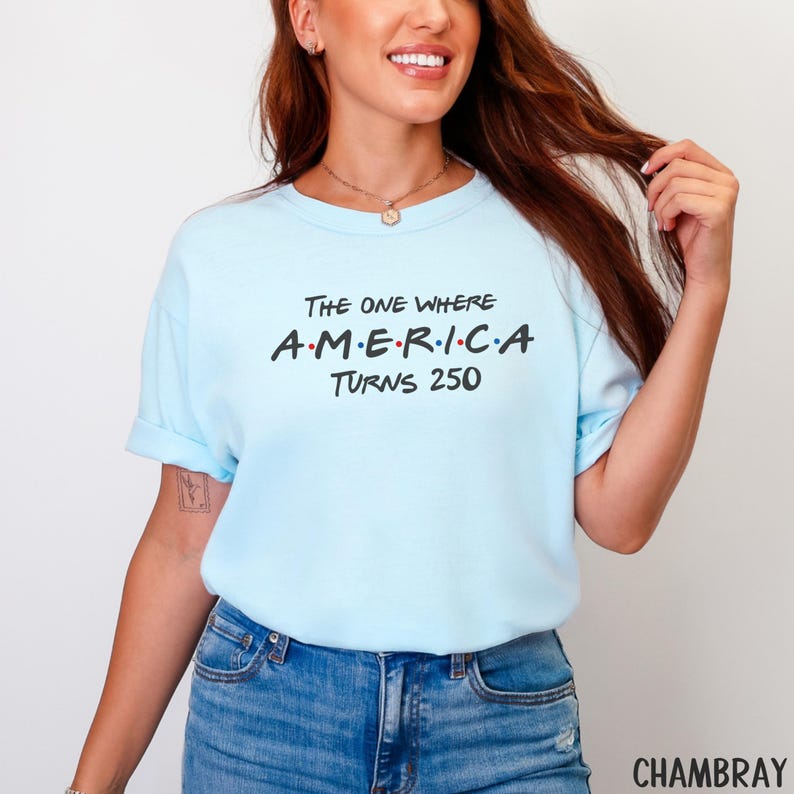 K&ouml;nnte beinhalten: Hellblaues T-Shirt mit dem Text "The One Where A.M.E.R.I.C.A Turns 250" in Schwarz und Rot. Das Shirt besteht aus weichem Material und hat hochgekrempelte &Auml;rmel. Ein l&auml;ssiger Stil.