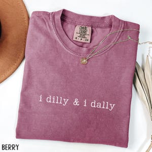 May include: Berry-colored t-shirt with the phrase "i dilly & i dally" in white. A gold necklace with a pendant rests on the shirt. The shirt has a crew neck and a tag that reads "Comfort Colors".