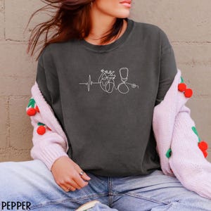 May include: Dark grey sweatshirt featuring a white line drawing of a heart, stethoscope, and heartbeat. The word "PEPPER" is printed in the lower left corner. The sweatshirt is worn with blue jeans and a pink cardigan.