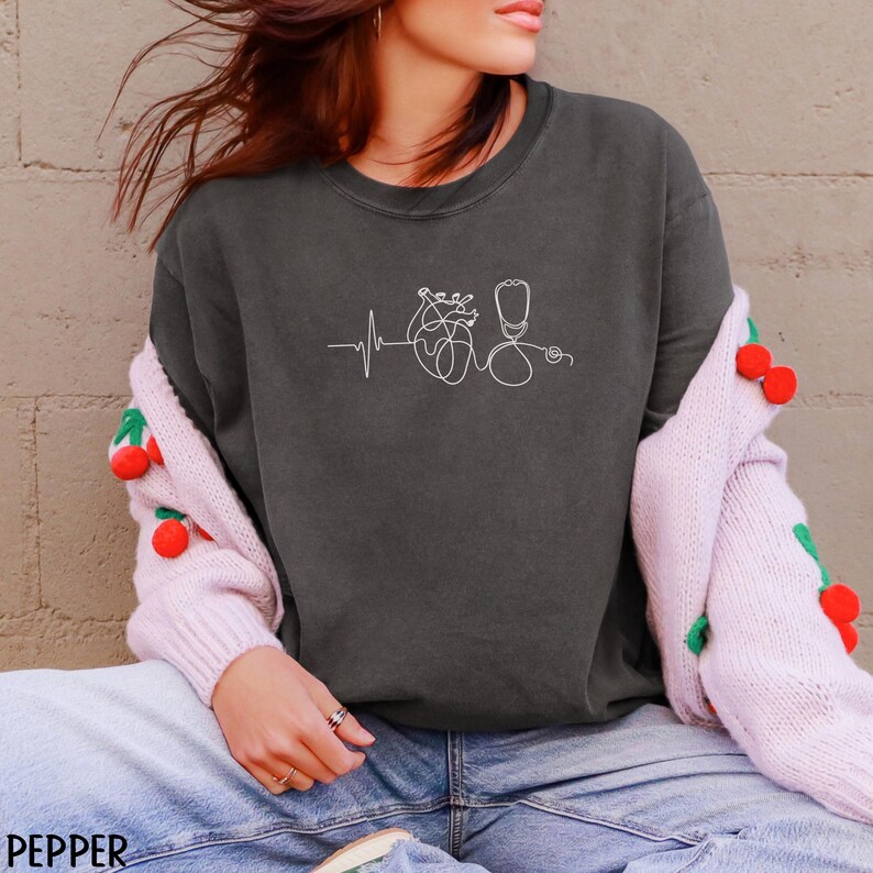 May include: Dark grey sweatshirt featuring a white line drawing of a heart, stethoscope, and heartbeat. The word "PEPPER" is printed in the lower left corner. The sweatshirt is worn with blue jeans and a pink cardigan.