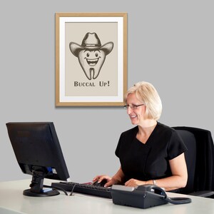 May include: A framed print with a cartoon tooth wearing a cowboy hat and the text "Buccal Up!" hanging on a gray wall. A woman is sitting at a desk in front of a computer.