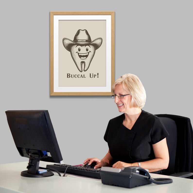 May include: A framed print with a cartoon tooth wearing a cowboy hat and the text "Buccal Up!" hanging on a gray wall. A woman is sitting at a desk in front of a computer.