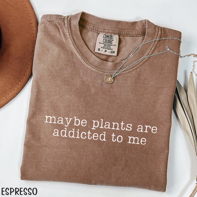 Może przedstawiać: Koszulka w kolorze espresso z białym napisem &bdquo;maybe plants are addicted to me&rdquo;. Srebrny naszyjnik z małą zawieszką jest zarzucony na koszulkę. Koszulka ma okrągły dekolt i kr&oacute;tkie rękawy.
