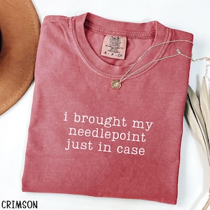 May include: A crimson short-sleeved t-shirt with a crew neckline and the white printed text "i brought my needlepoint just in case". The shirt features a Comfort Colors brand label at the inner neck and a delicate gold-toned necklace.