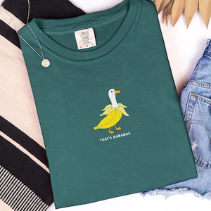May include: Teal Comfort Colors t-shirt with a graphic of a duck in a banana peel, with the text "THAT'S BANANAS". The shirt is next to a gold necklace and denim shorts.