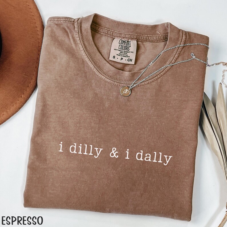 May include: Espresso-colored t-shirt with the phrase "i dilly & i dally" in white. The shirt has a crew neck and short sleeves. A silver necklace with a pendant is draped across the shirt. The shirt is made of a soft, comfortable material.