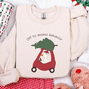 May include: Cream-colored sweatshirt with the phrase "Off to assess behavior" printed in brown. The graphic shows a goose wearing a Santa hat driving a red car with a Christmas tree on top. The sweatshirt has a crew neck.
