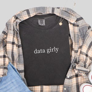 May include: A dark gray t-shirt with the words "data girly" in white script. The shirt is layered under a plaid button-down shirt in shades of gray, tan, and white. A gold necklace and denim jeans are also visible.