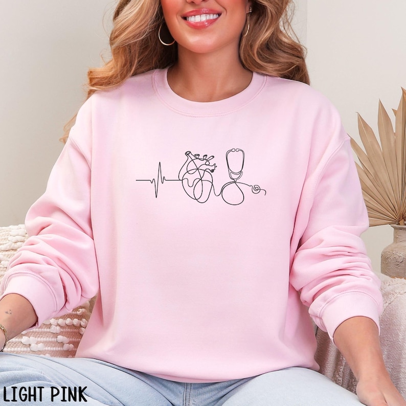 May include: Light pink sweatshirt featuring a minimalist line drawing of a heart, stethoscope, and heartbeat line. The text "LIGHT PINK" is at the bottom. The design is in black. The sweatshirt is a solid light pink color.