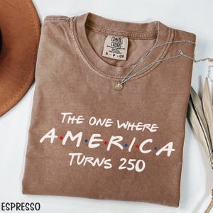 K&ouml;nnte beinhalten: Espressofarbenes T-Shirt mit dem Text "The One Where America Turns 250" in Wei&szlig;, Rot und Blau. Eine silberne Halskette mit Anh&auml;nger liegt auf dem Shirt. Das Shirt ist von der Marke Comfort Colors.