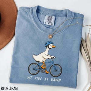 Puede incluir: Una camiseta azul de cuello redondo con un gráfico de un ganso blanco que lleva un casco de bicicleta azul y monta una bicicleta azul. El texto "WE RIDE AT DAWN" está impreso debajo del gráfico. La camiseta tiene una etiqueta de Comfort Colors.