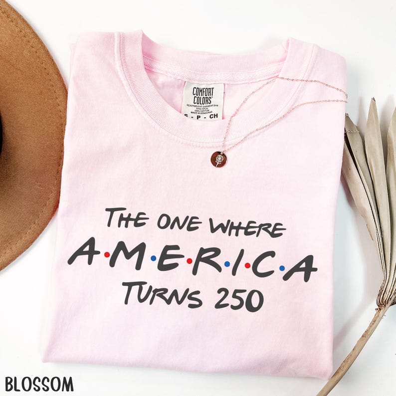 K&ouml;nnte beinhalten: Ein hellrosa T-Shirt mit dem Text "The One Where A.M.E.R.I.C.A. Turns 250" in Schwarz, Rot und Blau. Eine goldene Halskette mit Anh&auml;nger liegt auf dem Shirt. Das Wort "Blossom" steht unten links.