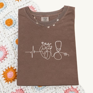 May include: A brown Comfort Colors t-shirt featuring a white line drawing of a heart, stethoscope, and heartbeat. A silver butterfly necklace is draped across the shirt. The shirt rests on a crochet blanket.