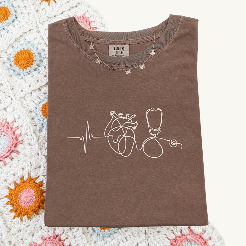 May include: A brown Comfort Colors t-shirt featuring a white line drawing of a heart, stethoscope, and heartbeat. A silver butterfly necklace is draped across the shirt. The shirt rests on a crochet blanket.