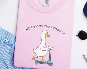 May include: A light pink t-shirt with the text "Off to observe behavior" and a cartoon goose riding a scooter. The goose is white with a yellow beak and feet. The scooter is light blue.