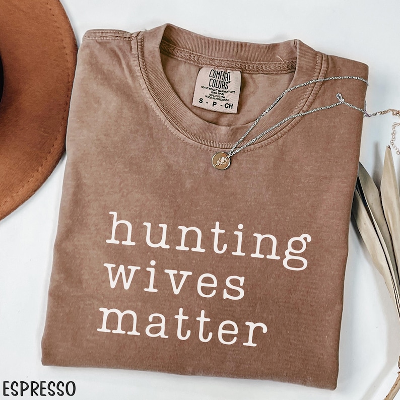 K&ouml;nnte beinhalten: Espressofarbenes T-Shirt mit dem Text "hunting wives matter" in wei&szlig;en Blockbuchstaben. Eine silberne Halskette mit Anh&auml;nger liegt auf dem Shirt. Das Shirt ist gefaltet und mit einem braunen Hut ausgestellt.