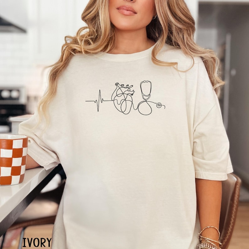 May include: Ivory-coloured t-shirt with a minimalist line drawing of a heart, stethoscope, and heartbeat line in black. The shirt has a relaxed fit and long sleeves. The word "IVORY" is printed at the bottom.