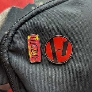May include: Two enamel pins on a black fabric background. The first pin is a red and yellow rectangle with the word "OLDZOY" written vertically. The second pin is a red circle with a black "V" inside.