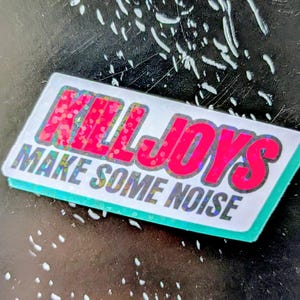 Peut inclure: Autocollant rectangulaire avec les mots "KILLJOYS MAKE SOME NOISE" en lettres rouges et pailletées argentées. L'autocollant a un fond blanc et une bordure turquoise, sur une surface sombre.