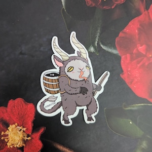 Puede incluir: Una pegatina con una criatura gris caricaturesca con cuernos, un barril en la espalda y un palo. La criatura tiene ojos amarillos y una lengua roja. La pegatina está sobre un fondo oscuro con flores rojas.