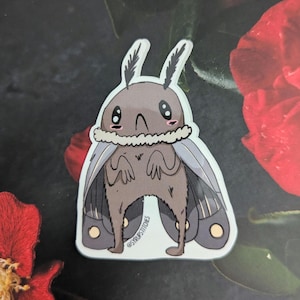 Puede incluir: Una pegatina con una polilla de dibujos animados con cuerpo marrón, alas grises y detalles blancos. La polilla tiene ojos grandes y una expresión linda. La pegatina tiene el texto "@syrupstitches" en la parte inferior.