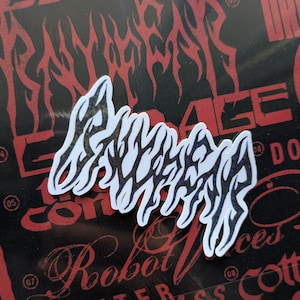 May include: A white sticker featuring black tribal-style lettering, set against a black background with red graphic elements. The sticker's design is a stylized word, potentially a brand name.