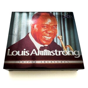 May include: A CD box set featuring a portrait of Louis Armstrong, smiling and holding a trumpet. The box set is titled "Louis Armstrong Triple Treasures" and includes a "3 CD" label.