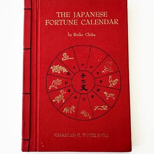 Könnte beinhalten: Ein rotes Buch mit dem Titel "THE JAPANESE FORTUNE CALENDAR" von Reiko Chiba. Das Cover zeigt ein goldfarbenes kreisförmiges Design mit Tierillustrationen des Tierkreises und japanischen Schriftzeichen. Das Buch ist mit schwarzem Faden gebunden.