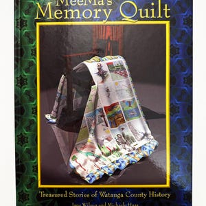 May include: A book titled "MeeMa's Memory Quilt" with a colorful quilt draped over a chair on the cover. The quilt features various illustrated scenes. The book's cover has a gold border and the text "Treasured Stories of Watauga County History".