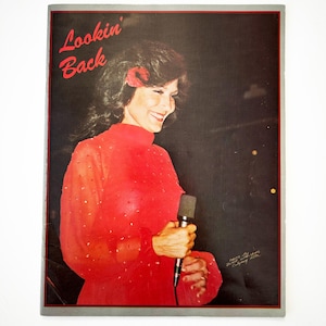 May include: A framed photograph of a woman in a red dress holding a microphone. The text "Lookin' Back" is in red script in the upper left corner. The background is dark, and the woman is smiling.